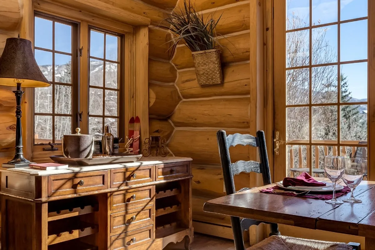 A sunlit wooden dining table set for a meal, complemented by wine glasses, a lamp, and large windows offering peaceful views. The setting is intimate yet inviting, perfect for group dinners or casual gatherings.
