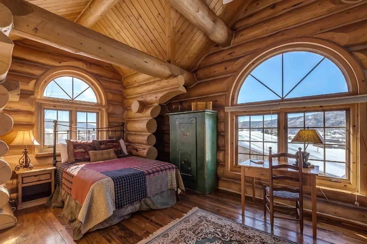 A spacious bedroom filled with natural light from large windows, featuring a bed with colorful bedding, an antique green cabinet, and a wooden chair and table set by the window. The room exudes warmth and provides an ideal setting for relaxing or enjoying the scenery.