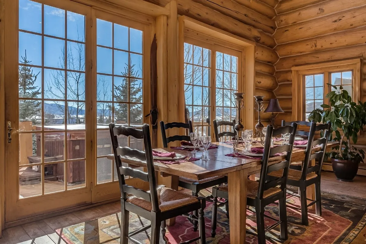 A spacious dining area with a large wooden table is surrounded by windows and accented by exposed beams and a decorative plant. The setting is ideal for communal meals or festive celebrations, bathed in natural light that highlights the welcoming, rustic elegance of the space.