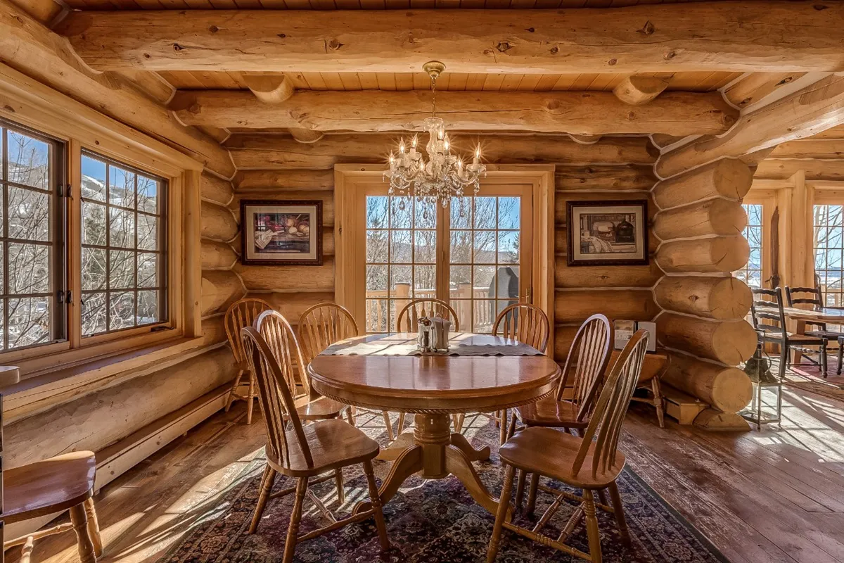 A spacious dining area with a wooden table, elegant chandelier, large windows, and log walls. The room is filled with natural light, suggesting a connection to mountain views and outdoor beauty. The setting is warm and communal, ideal for family meals and post-adventure relaxation.