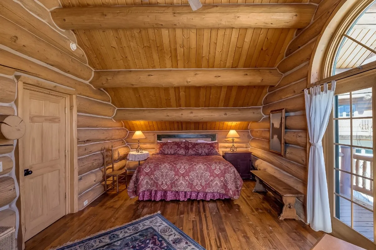 A bedroom with log walls, ornate bedding, a large window overlooking nature, and wooden furniture. The warm tones and textures create a welcoming environment, inviting relaxation and restful sleep.