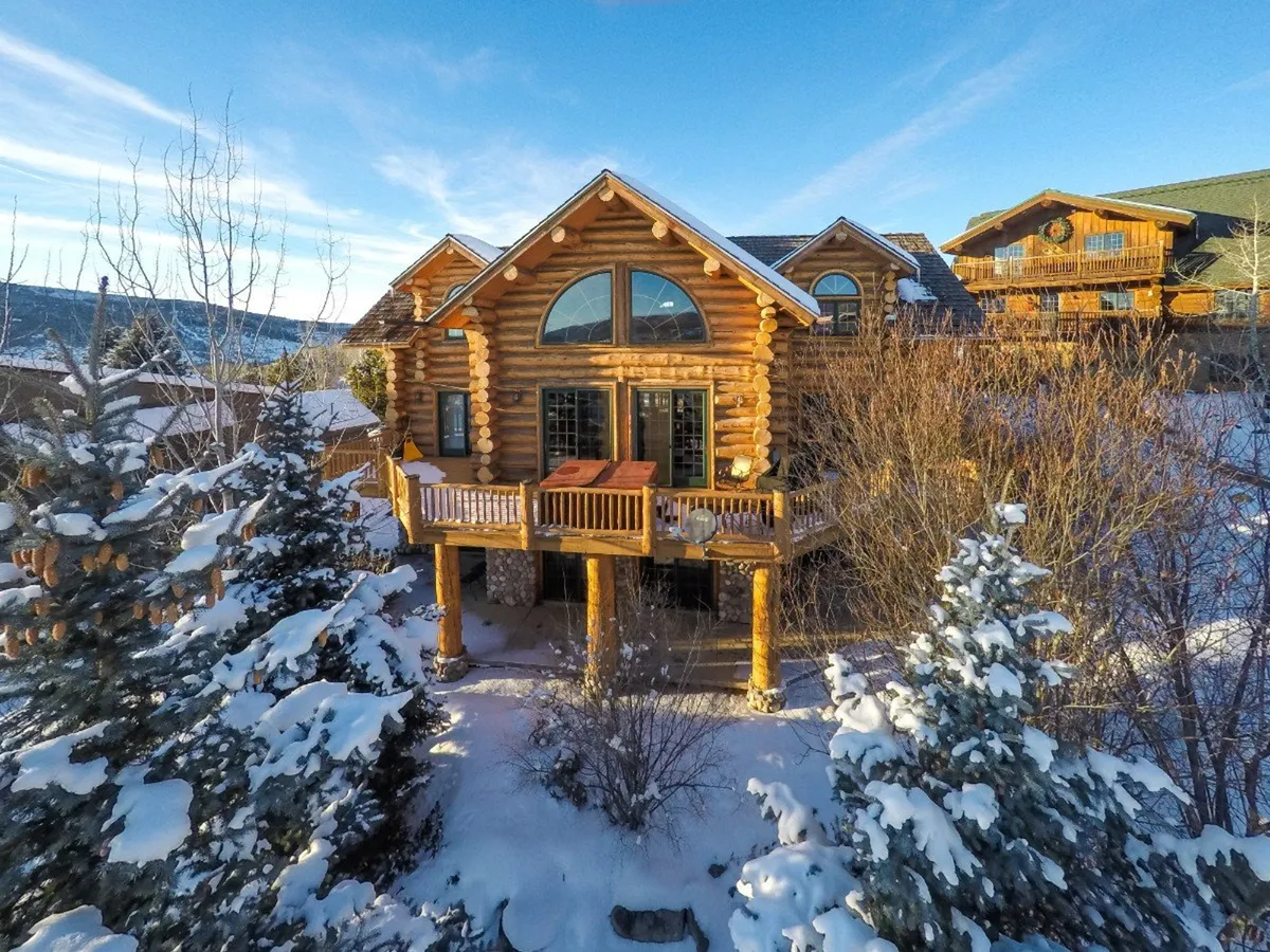 This exterior image features the log cabin structure with large windows, a deck equipped with a hot tub, and a backdrop of snow-covered trees. The scene suggests both winter adventure and cozy relaxation, with easy access to outdoor fun through the visible trails and inviting spaces for family gatherings.