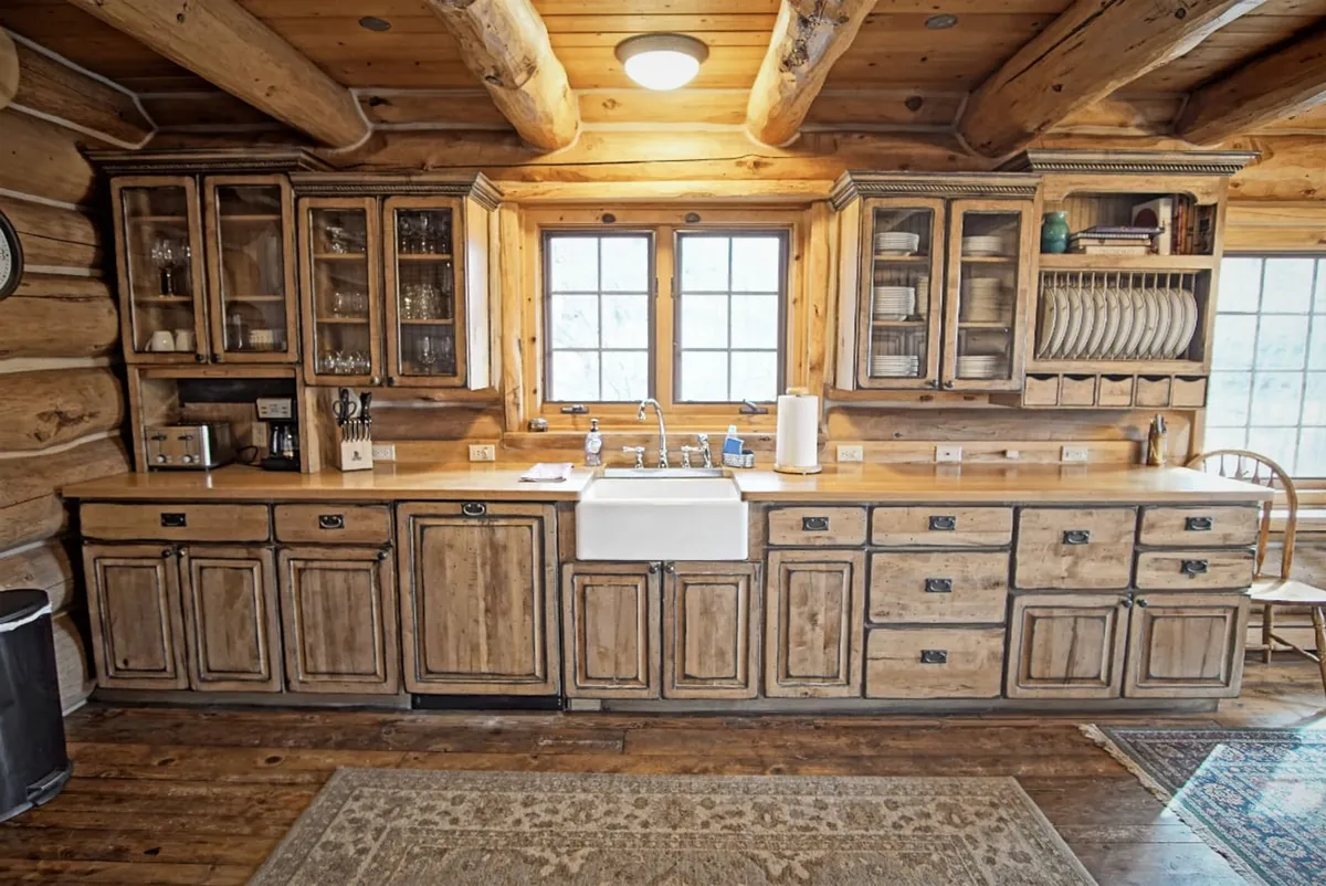 This image features a warm, inviting kitchen with wooden cabinetry, a farmhouse sink, large windows that bring in ample natural light, and a dining chair beside a cozy rug. The expansive windows offer a glimpse of the outdoors, seamlessly connecting the interior to the exterior and evoking a sense of summer gatherings and barbecue preparation.