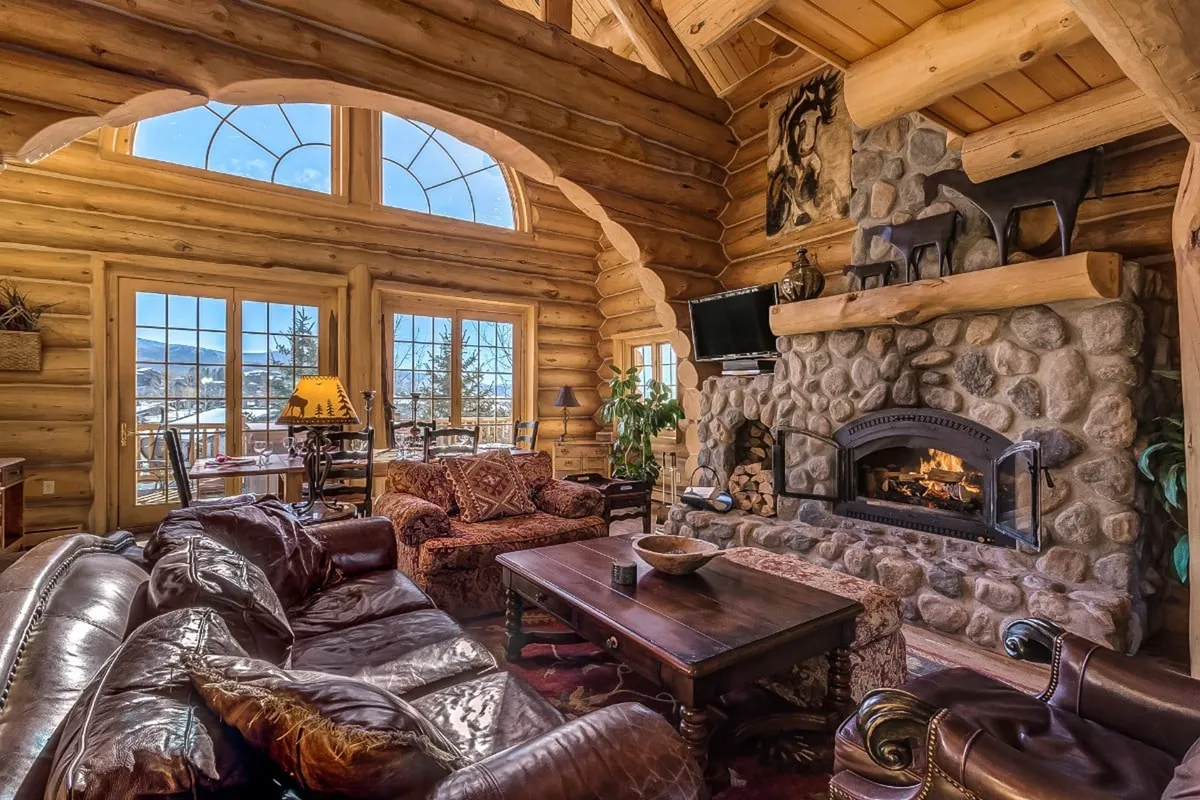 This interior image showcases the main living room centered around a large stone fireplace, with sturdy wooden beams overhead and comfortable leather sofas arranged for conversation. The space is warmly lit, and the arrangement encourages both group gatherings and intimate moments like reading or relaxing by the fire. Large windows in the background allow sunlight to pour in, highlighting the welcoming atmosphere.
