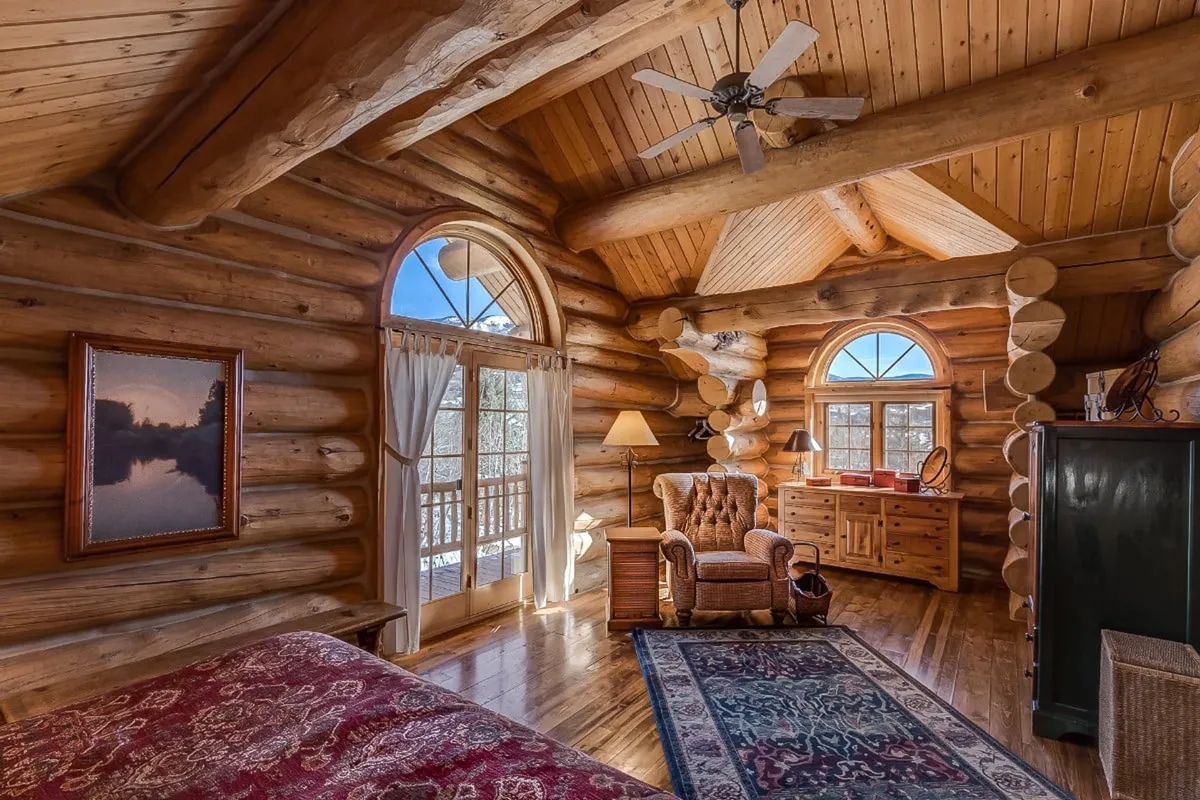 A tranquil reading nook with log walls, large windows, antique furniture, and a soft area rug. The space is designed for quiet moments, with chairs perfectly placed for reading or enjoying the outdoor view. Warm, golden light creates a sense of peaceful privacy within the lodge.