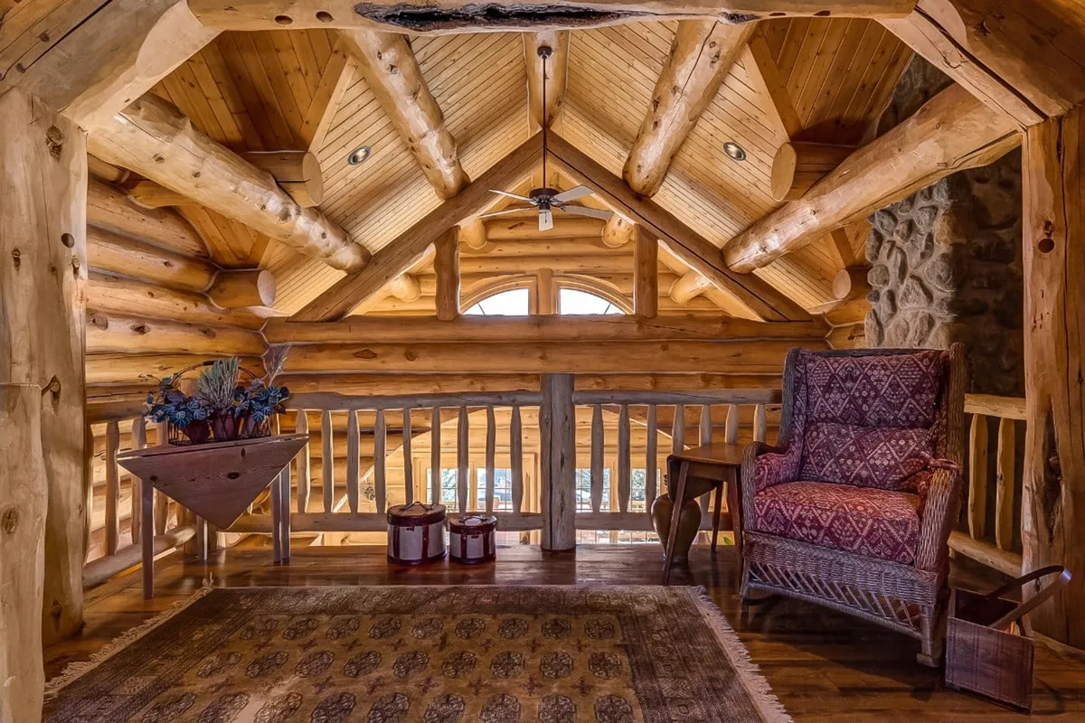 A warm, tranquil living area featuring rustic wooden beams, large windows that invite natural light, a plush armchair by the window, a decorative rug, and a rustic table. The arrangement creates a cozy space that encourages relaxing and enjoying views beyond the home.