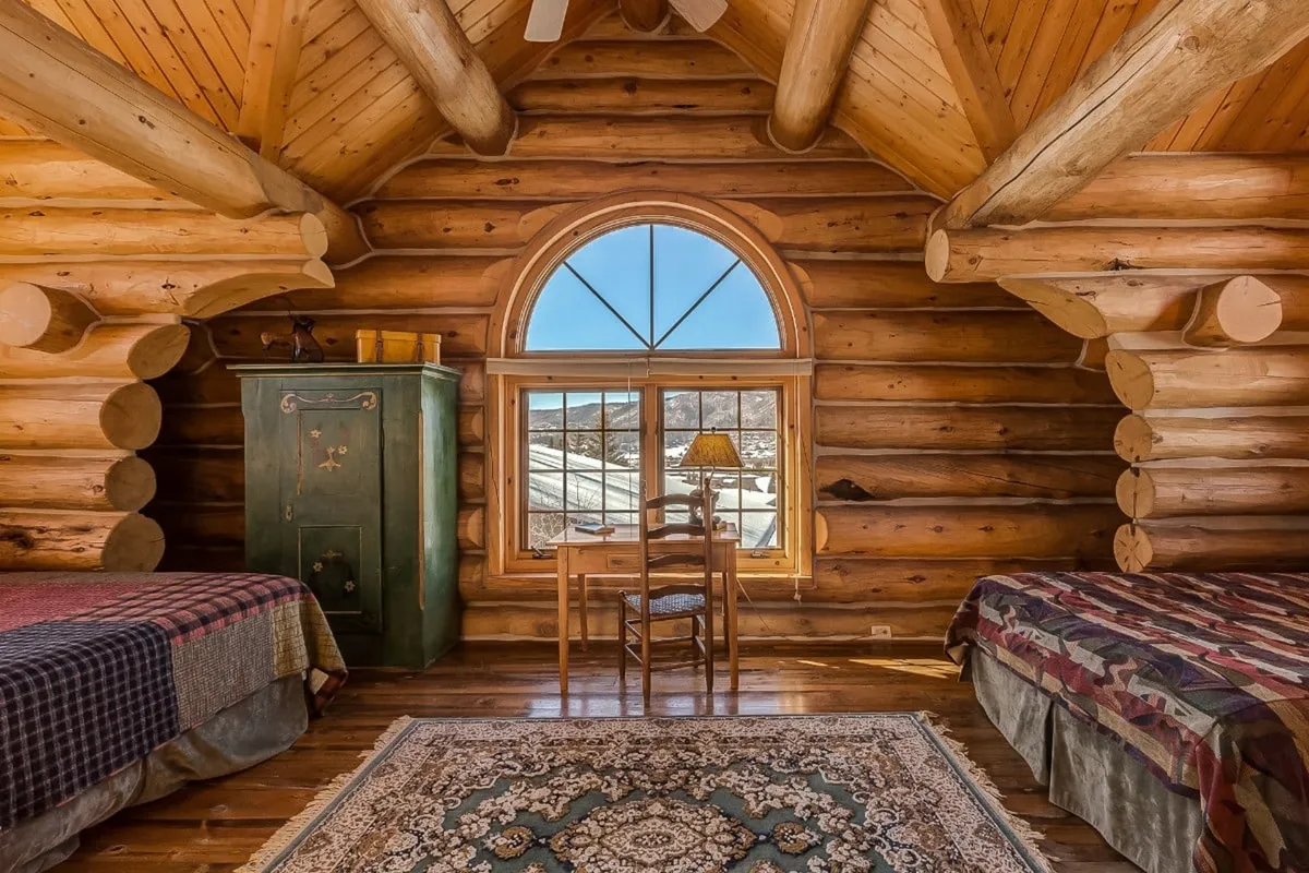 A tranquil bedroom with log walls, an arched window bathing the space in natural light, cozy wooden furniture, and a plush rug. The atmosphere is serene, ideal for quiet moments and restful sleep.