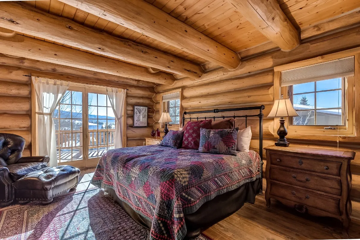 A warmly lit bedroom featuring a bed with a patterned quilt, a leather armchair perfect for reading, a wooden dresser, and large windows dressed with curtains that frame the view outside. The decor is cozy and inviting, with soft lighting enhancing the restful ambiance.