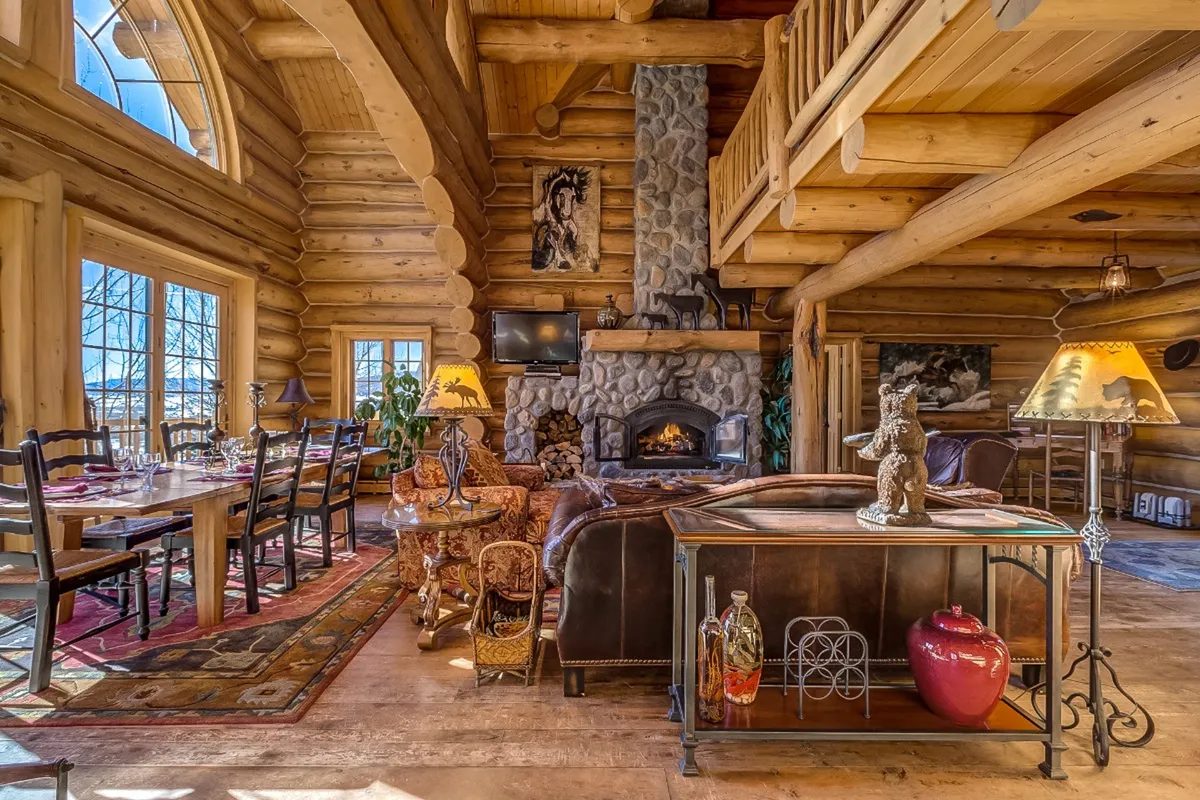 This inviting interior scene showcases soaring wooden beams, a grand stone fireplace as the centerpiece, a spacious dining table perfect for gatherings, and large windows that flood the space with natural light. The room exudes mountain lodge charm with tasteful western decor and cozy seating, inviting guests to relax and share memorable moments together.