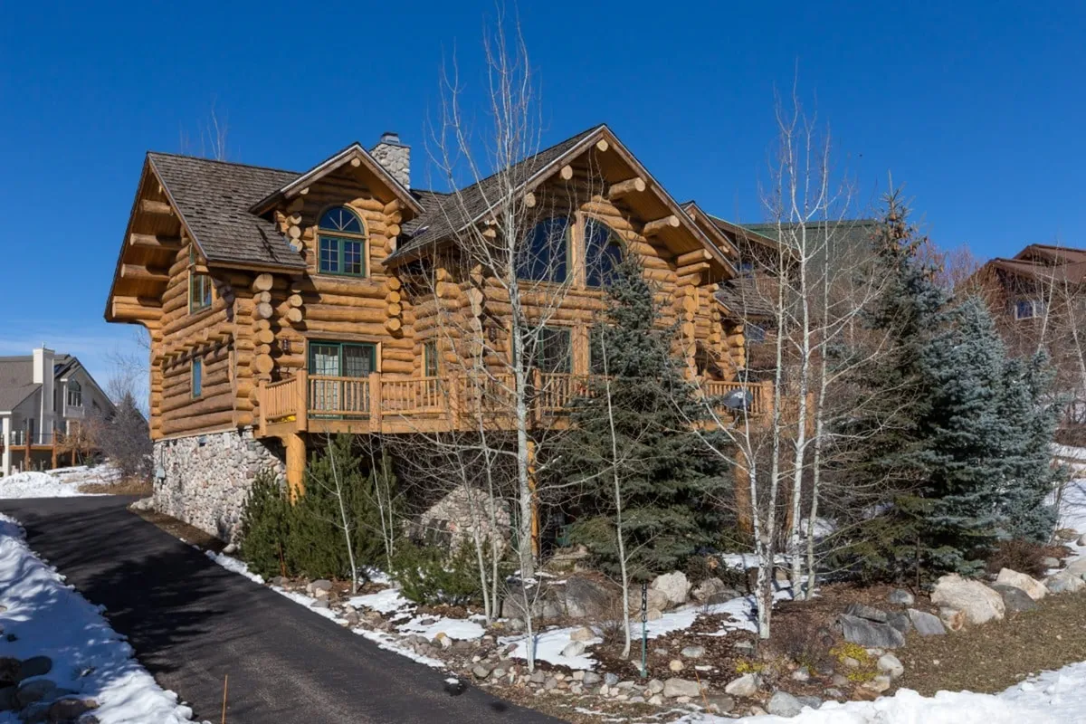 The cabin's exterior features large windows, inviting log construction, and stone accents, all surrounded by natural landscape. The scene hints at outdoor gathering spaces and the seamless transition from indoor comfort to outdoor exploration.