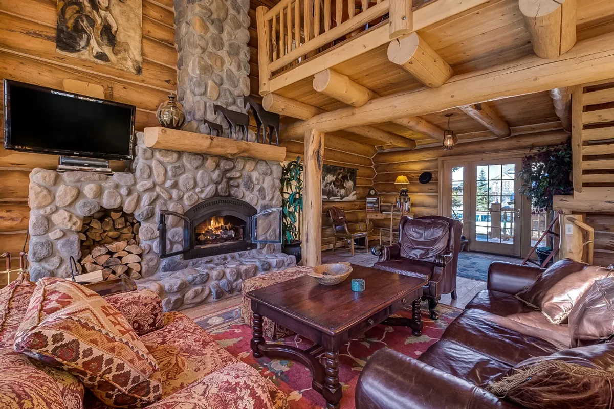 This warm, homely living area features a stone fireplace, leather armchairs, a wooden coffee table, and large windows. Decorative pillows and seasonal touches add to the inviting mood, while the seating arrangement supports both group gatherings and private relaxation. Sunlight streams in, reinforcing the link between cozy interiors and the surrounding landscape.