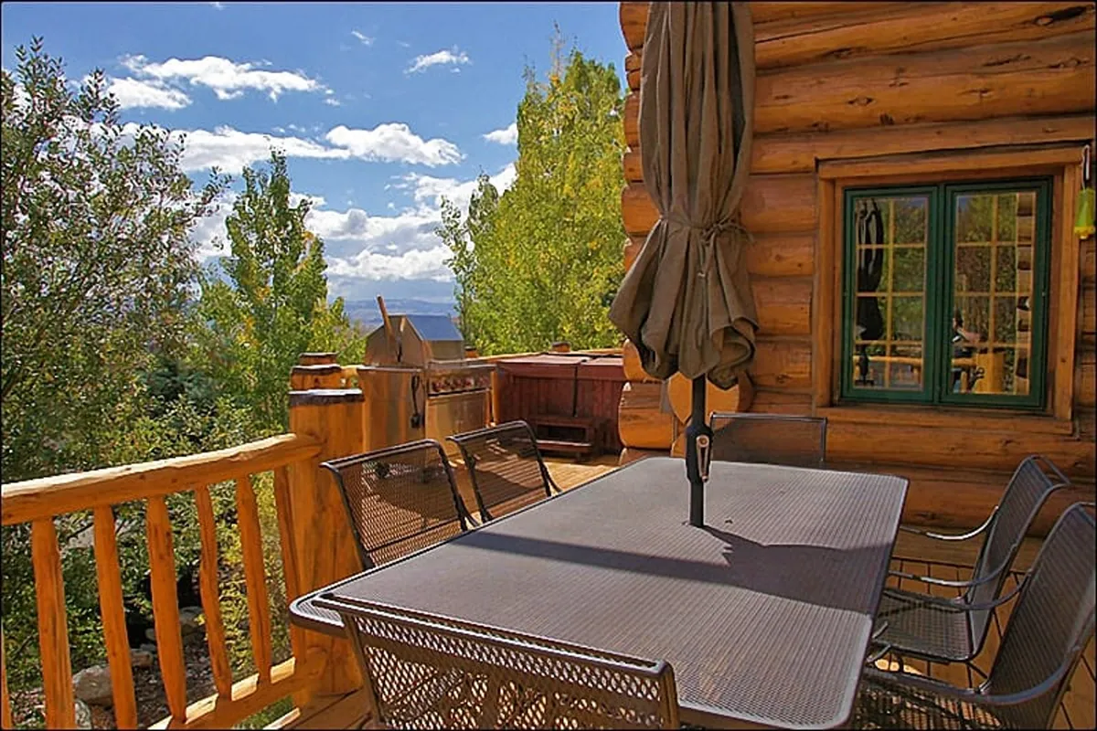 An inviting outdoor dining scene featuring a log cabin exterior, a large dining table under an umbrella, green surroundings, and distant mountain views. The space is set for al fresco meals, barbecues, and relaxed socializing with nature as a backdrop.